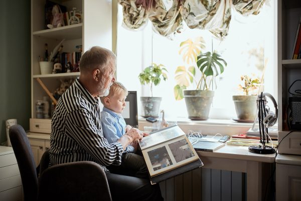 Senior man looking at photo almbum with male toddler in front of a window Senior male parent looking at photo memories album with future generation male toddler in front of a window